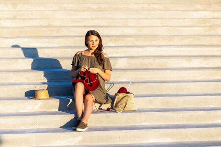 A young woman sitting In a deserted place cityscape, on the steps and knitting a sweater with knitting needles on a summer day. Get Creative everywhere during quarantineの写真素材