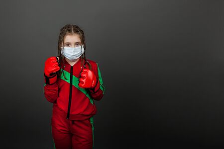 Cute young girl boxer in a medical mask and boxing gloves is standing in a boxing rack.の写真素材