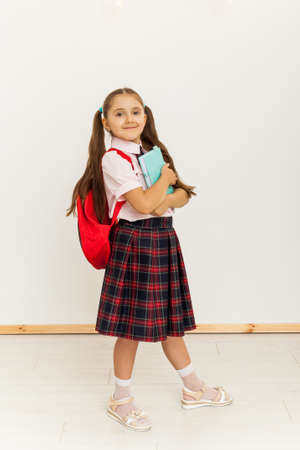 Full length portrait of a schoolgirl in uniform with backpack standing on white background and holding bookの写真素材