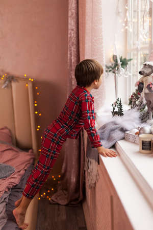 Cute Curious boy in plaid pajama, looking through a window, waiting impatiently for Santa and giftsの写真素材