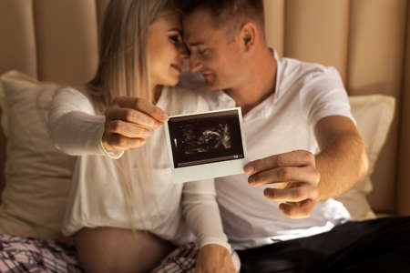 Happy parents holding baby ultrasound photo smiling at each other at home. Focus on ultrasound image