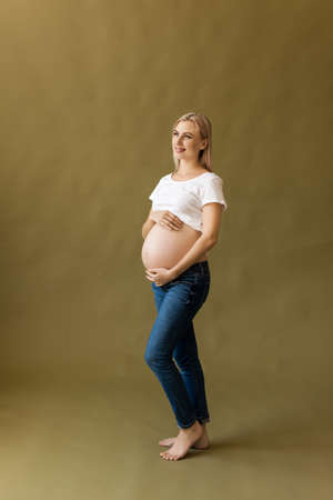 Full length portrait of pregnant woman touching her belly on beige background. Pregnancy, maternity, preparation and expectation conceptの写真素材