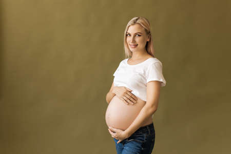 Pregnant happy Woman touching her belly. Pregnant mother portrait, caressing her belly and smiling. Healthy Pregnancy concept, blond expectant female on beige backgroundの写真素材
