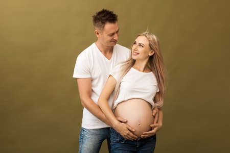 Image of pregnant smiling couple on beige background. Husband touching his wife belly with hands
