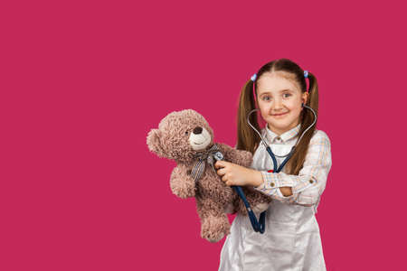 Pretty preschool child girl wear white medical uniform listening a bears heart with a stethoscope a...