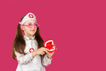 Little cool doctor dentist girl in a medical suit with a toy jaw and Toothbrush on pink backgroundの写真素材