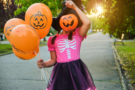 Funny Halloween kid girl carnival costumes hiding face with candy bucket outdoors. Portrait smiling girl in witch hat with orange balloons.の写真素材