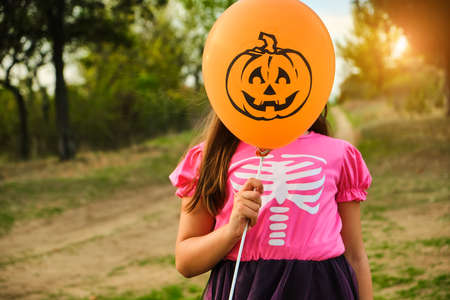 Funny Halloween kid girl in carnival costumes Hiding her face behind an orange balloon outdoors.の写真素材