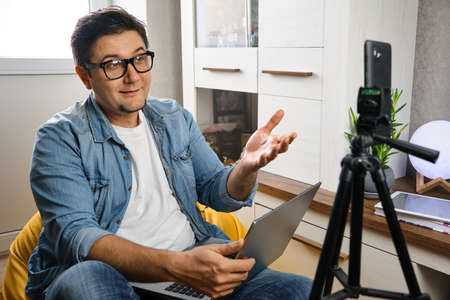 Hand free. Middle-aged male sitting at home before smartphone fixed on tripod. Video blogger...