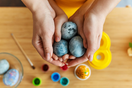 Mother and her kid hands holding painting Easter eggs. Female and child hand with egg over the table with paints and brushes. Happy family preparing for Easter. Close up, selective focusの写真素材