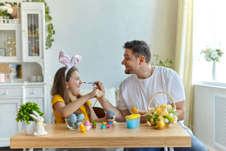 dad and daughter stain each other's faces with blue paint for painting eggs. on the table is a basket with Easter eggs and paints.の写真素材