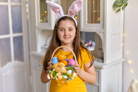 Cute little child wearing bunny ears on Easter day. Girl holding basket with painted eggs in sunny spring day indoor.の写真素材