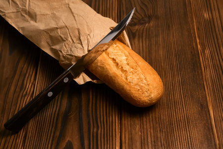 Fresh baguette in a craft package bread knife on brown wooden background