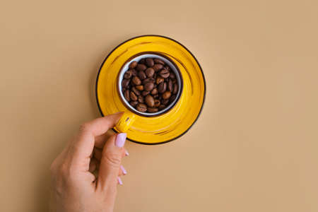 Arabica coffee beans in a bright designer yellow cup with saucer in female hand on a pastel beige paper background. top view, copy spaceの写真素材