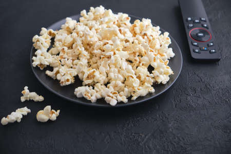 Black plate of popcorn with salted, remote control TV on a black table. View from above. Leisure. Watch TV.の写真素材