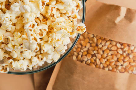 Close up of Cooked Popcorn in glass bowl and corn grains in a paper box on a beige background....