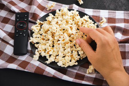 Close up of female hand taking popcorn from black plate watching movie at home.