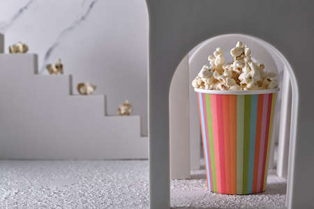 Striped paper cup with popcorn in a decorative arch and steps with popcorn, trendy geometric shapes. Cinema and snack creative food concept.の写真素材