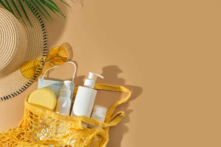 palm, bucket sun hat, SPF sun protection cream in string bag, sunglasses and surgical mask on beige...