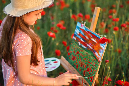 Little girl is standing in the field of red poppies and painting on the canvas placed on a drawing stand. Be a part of learning outside of the school in the nature park. Creative hobbyの写真素材