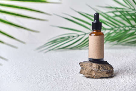 Summer mockup concept for product presentation cosmetic bottle on stone platform. dropper brown glass bottle with blank label on white background with green palm leavesの写真素材