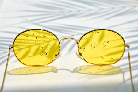 Close up of Sunglasses with yellow glasses with water drops lie on a water background. The shadow of a palm tree, copy space. Summer travel and vacation conceptの写真素材