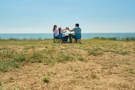 Happy family enjoying time at capmsite outdoors. Parents and their kid sitting together. On a cliff above the sea, they sit on tourist chairs, have a snack, communicate, enjoy their rest.の写真素材