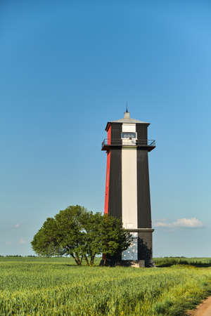 Beautiful landscape with Black and white lighthouse in a wheat green field and a lonely tree against...
