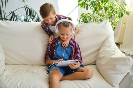 Cute brother and sister with headphones enjoying tablet at home. family, children, technology and home conceptの写真素材
