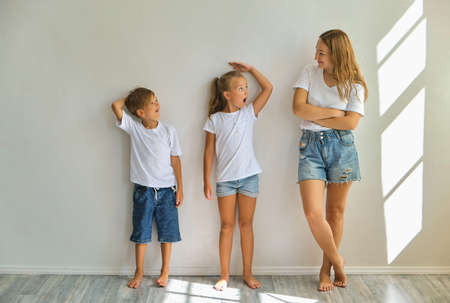 concept of the family. mother measures growth of kids to a daughter and son near light gray wall...