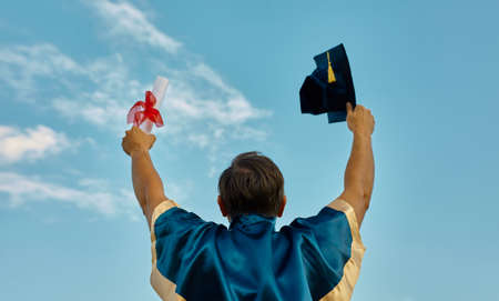 excited graduate student in gown with risen hands holding diploma over blue skyの写真素材