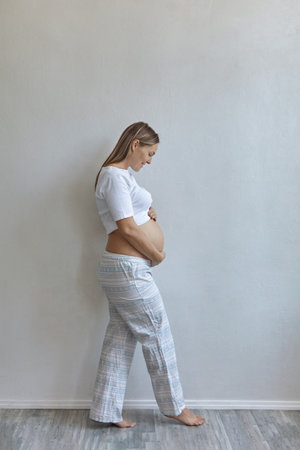 Beautiful pregnant woman in pajamas stands against the background of a white wall at home