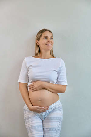 Beautiful pregnant woman in pajamas stands against the background of a white wall at home