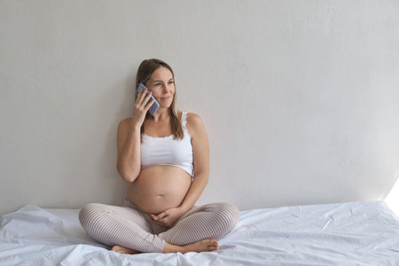Smiling pregnant woman which sitting on bed and using smartphone in homeの写真素材