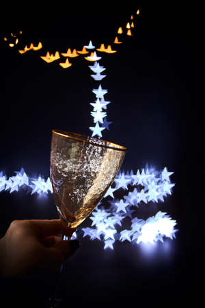 Golden festive glass in hand on a dark background and magic bokeh in the form of stars pouring into the glass. Christmas background, place for text.の写真素材