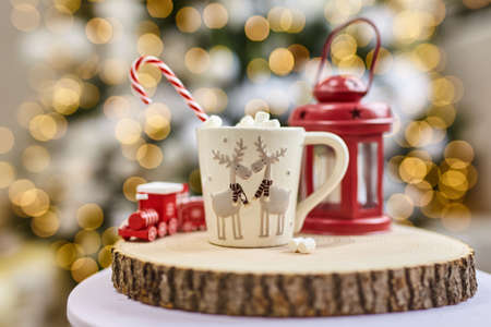 Fairy lights background, hot cocoa with marshmallows, white cup with deer on a wooden cut and red lantern and children's train at home during Christmas and New Year holidays. Soft focus bokeh.の写真素材