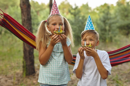 Children eating birthday cakes with burning candles. Kids party decoration and food. Boy and girl celebrating birthday in the garden with hammock. Kids with sweets.の写真素材