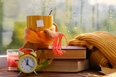 Hello Autumn. Yellow alarm clock, cup of tea on books near the window, it's raining. Mock up for tea designの写真素材