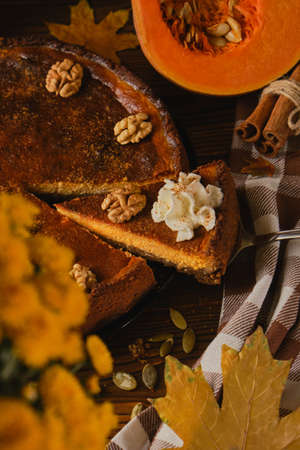 Fresh homemade pumpkin cheesecake with whipped cream and walnuts, with ingredients and autumn flowers on the wooden tableの写真素材