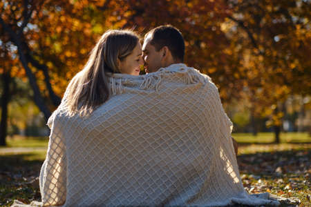 Beautiful couple man woman in love. Man and woman wrapped in beige blanket hugging together in park on autumn fall sunny day. Authentic real people.の写真素材