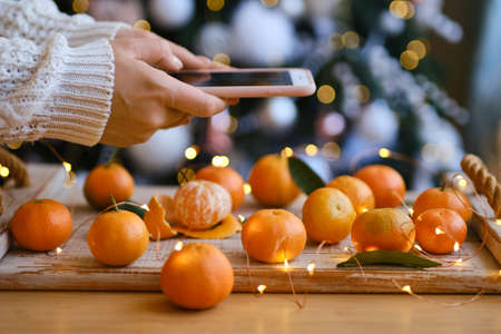 woman in knitted sweater makes a photo on smartphone of Christmas composition with mandarines for festive mood, fir tree decoration behind. holiday aroma. Selective focusの写真素材
