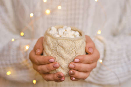 Woman in warm white sweater is holding white latte mug with marshmallow in hands. Mockup for winter gifts design with garland lights. Nail design. Winter soft pink color manicure. Christmas or winter compositionの写真素材