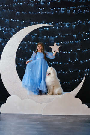 Beautiful little girl in light blue dress, stay on the moon in clouds with white Samoyed dog. Fabulous photo in studioの写真素材