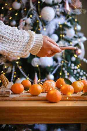 woman in knitted sweater makes a photo on smartphone of Christmas composition with mandarines for festive mood, fir tree decoration behind. holiday aroma. Selective focusの写真素材