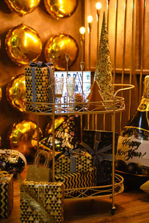 Golden serving table on wheels table with Christmas decor, many gift boxes and lightbox with the words Happy new Year, against wall with round golden balloonsの写真素材