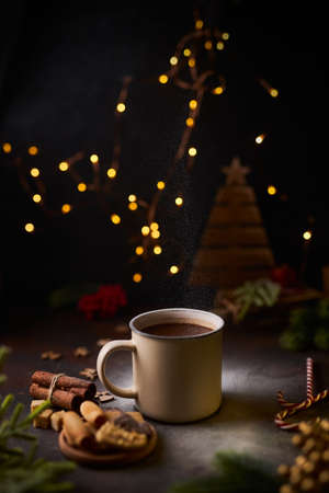 Mug of hot chocolate With a cloud of powder like snow, surrounded by Christmas decorations, green spruce branches, cinnamon, coockies and candy canes, against a dark background. Xmas moody.の写真素材
