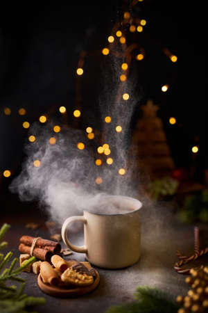 Mug of hot chocolate With a cloud of powder like snow, surrounded by Christmas decorations, green spruce branches, cinnamon, coockies and candy canes, against a dark background. Xmas moody.の写真素材