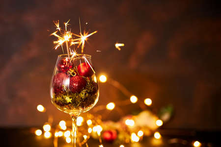 Happy New Year. Burning bengal firework in wine glass with red Christmas balls on background of golden bokeh lights. Creative aesthetic moment. Christmas atmosphere and mood at night. Selective focusの写真素材