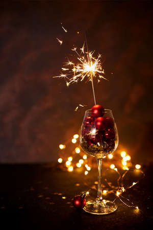 Happy New Year. Burning bengal firework in wine glass with red Christmas balls on background of golden bokeh lights. Creative aesthetic moment. Christmas atmosphere and mood at night. Selective focusの写真素材