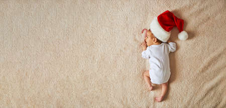 Little sleeping newborn baby, wearing Santa hat and white blank bodysuit, lies on fur blanket with garland. Top above view, banner size with copy space. x-mas home indoorsの写真素材